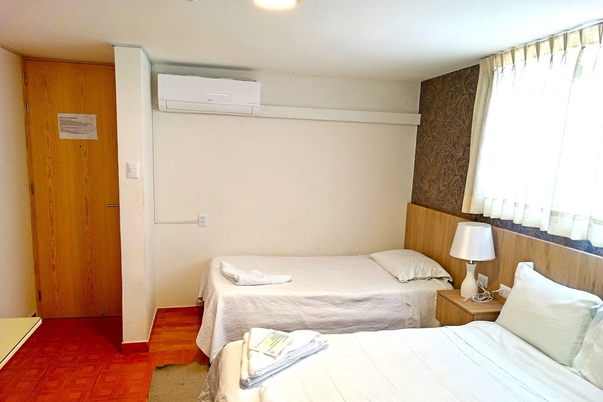 A hotel room with two single beds, a wooden wardrobe, an air conditioning unit, a bedside table with a lamp, and a window with curtains.