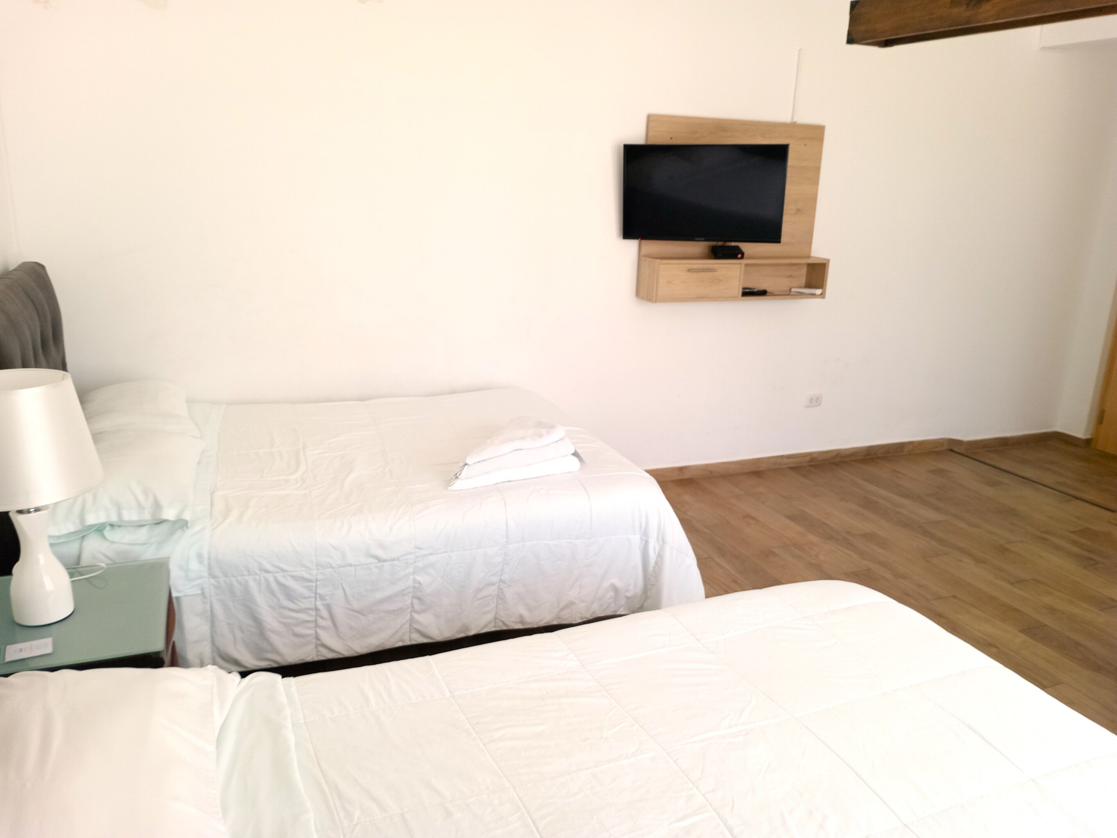 A hotel room with two beds, a TV mounted on the wall, a bedside table with a lamp, and a wooden floor.