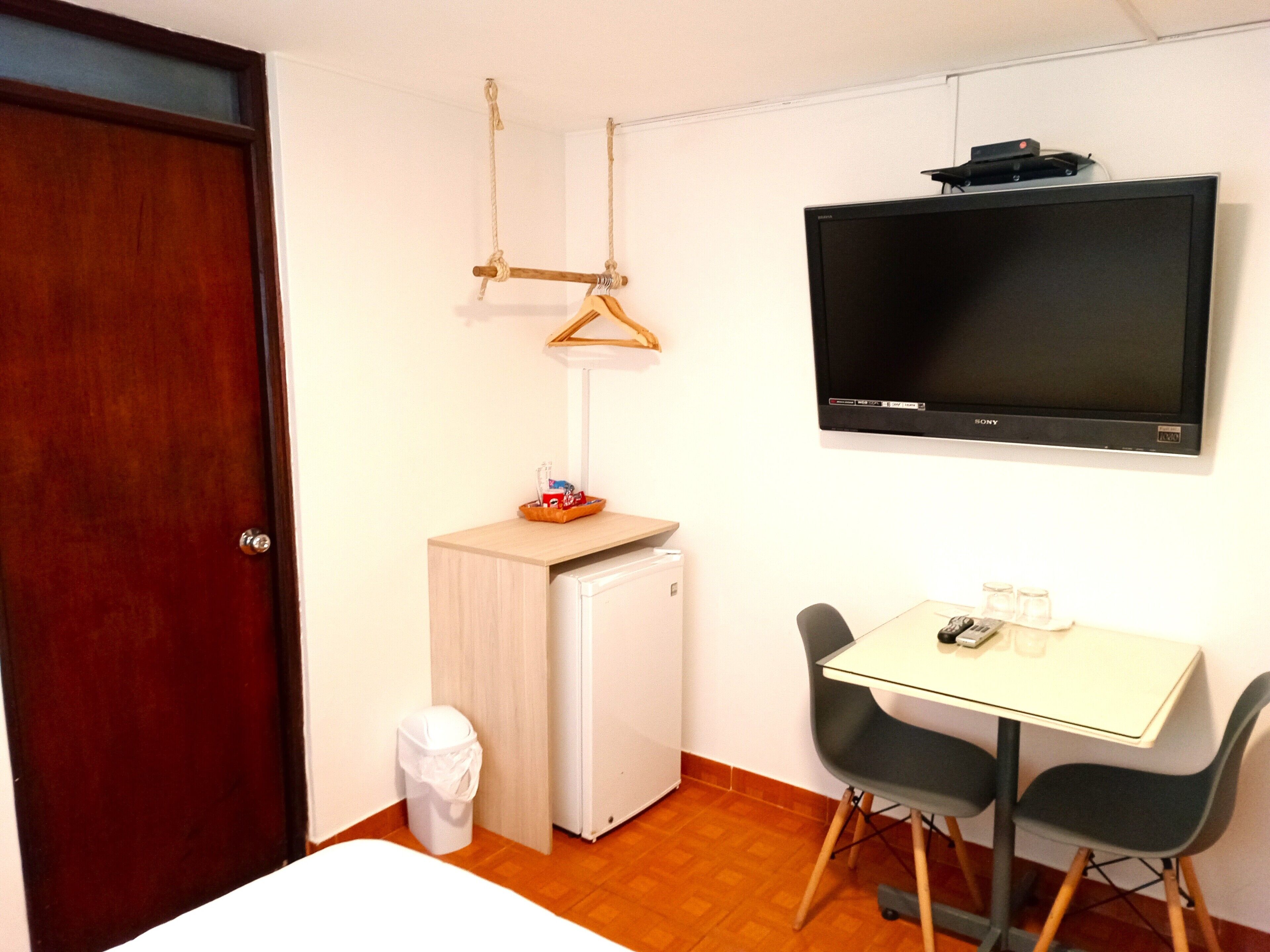 A small room with a bed, a desk with two chairs, a TV, a small fridge, and a trash bin.