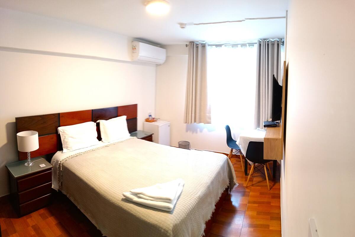 A hotel room with a bed, bedside table, lamp, desk, chair, and a window with curtains.