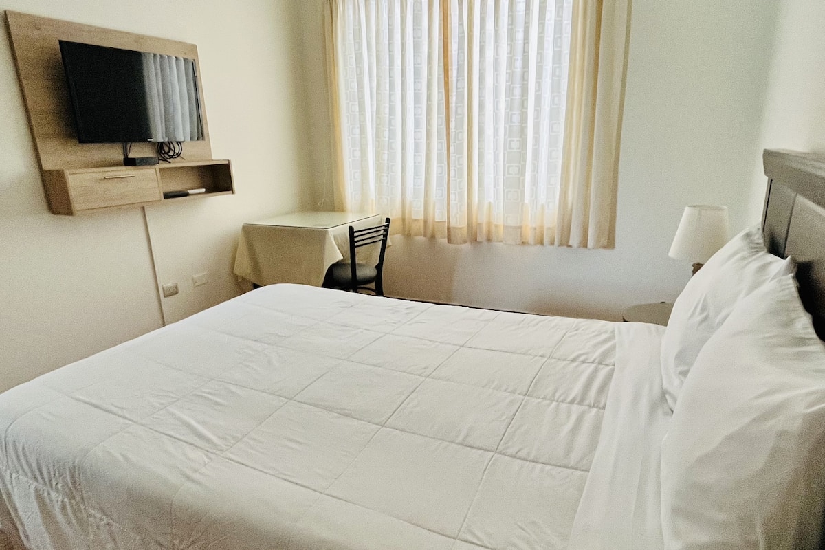 A hotel room with a large bed, a television mounted on the wall, a small table, and a chair.