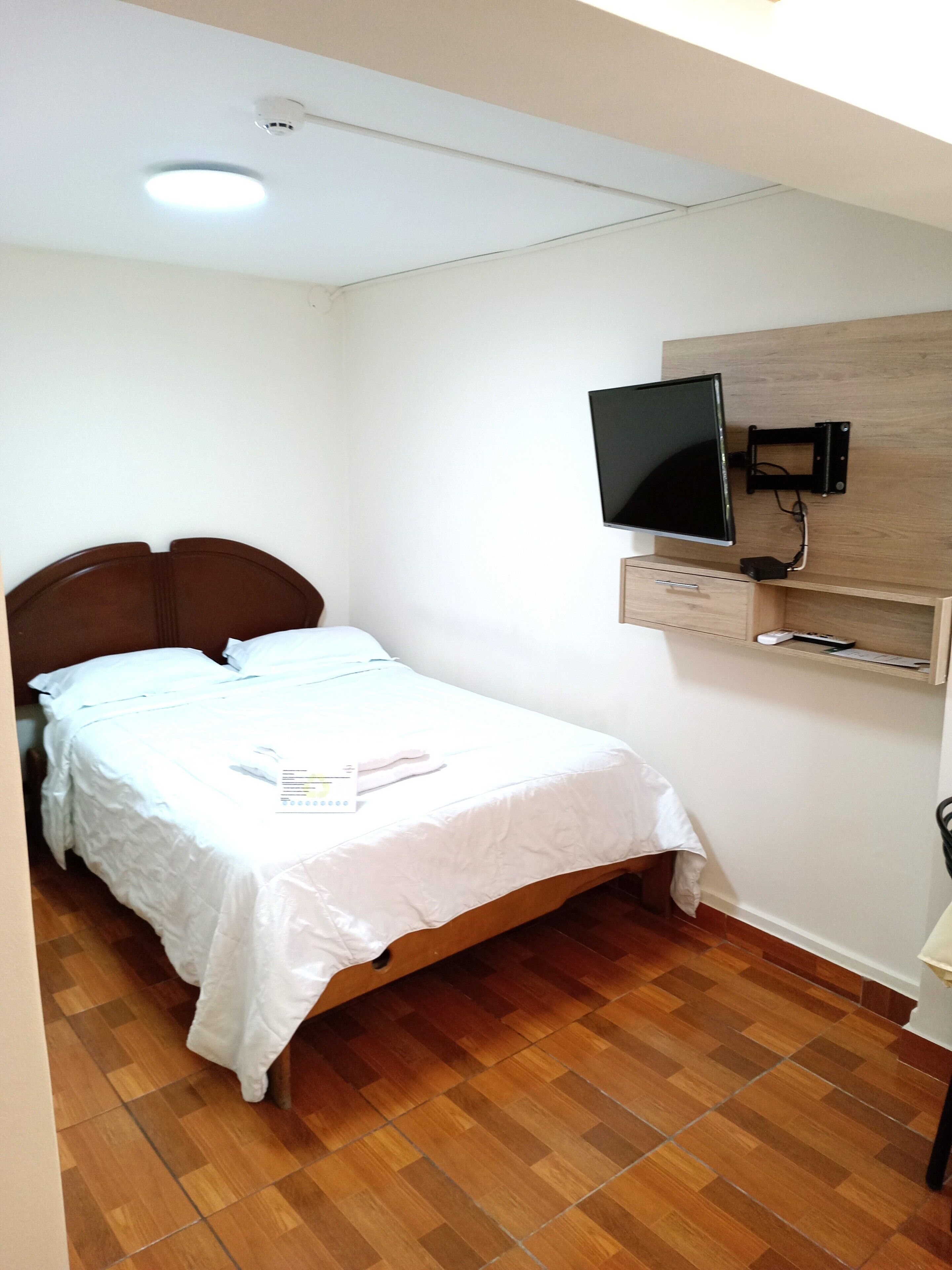 A single bed with a wooden headboard, a flat-screen TV mounted on a wooden panel above it, and a small wooden cabinet below.