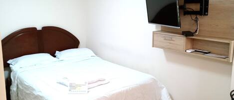 A single bed with a wooden headboard, a flat-screen TV mounted on a wooden panel above it, and a small wooden cabinet below.