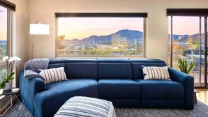 A modern living room with a large sectional sofa, a view of a cityscape and mountains through large windows, and a small side table with a lamp and a potted plant. - “Windmill House” -Mountain/City views, super close to great hiking trails (Phoenix)