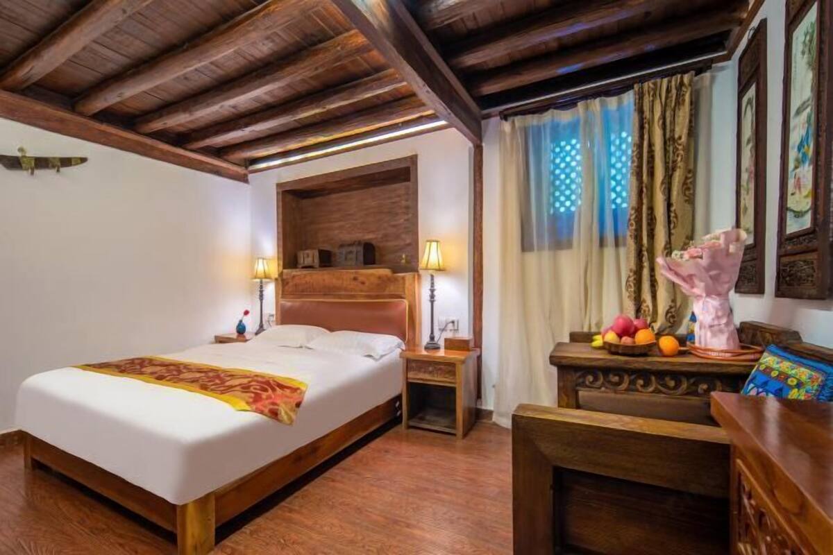 Quarto de hotel com cama de madeira, mesa de cabeceira, escrivaninha com luminária, janela com cortinas e parede com quadros.