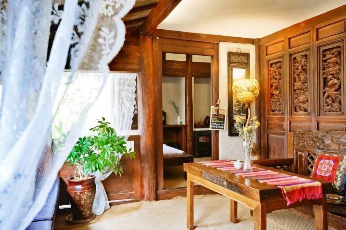 Quarto tradicional com mobiliário de madeira, uma mesa com toalha, uma planta em vaso e um painel de parede entalhado.