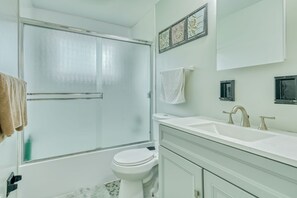 A bathroom with a glass shower door, a toilet, a sink with two faucets, and a mirror with decorative wall art.