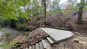 Uma trilha de pedra leva a um deck de madeira com cadeiras, cercado por uma área arborizada e um rio.