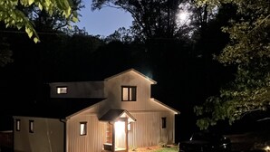 Uma casa de dois andares com varanda frontal e uma pequena garagem anexa, cercada por árvores e um caminho de acesso de cascalho.