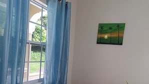 En beige sofa i et rum med et vindue med blå gardiner, et billede af en solnedgang med palmer og en potteplante.