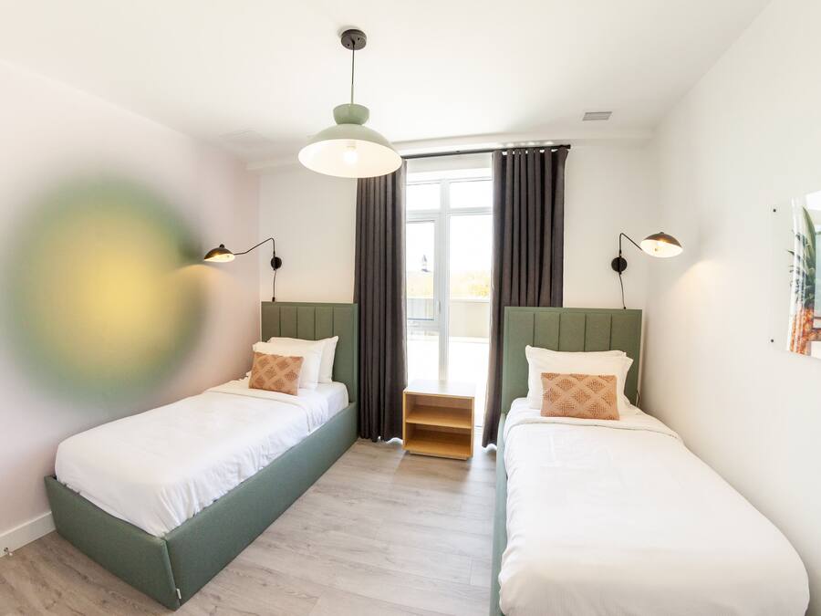 Una habitación de hotel moderna con dos camas individuales, una mesita de noche de madera, un cabecero verde y un ventanal con cortinas.