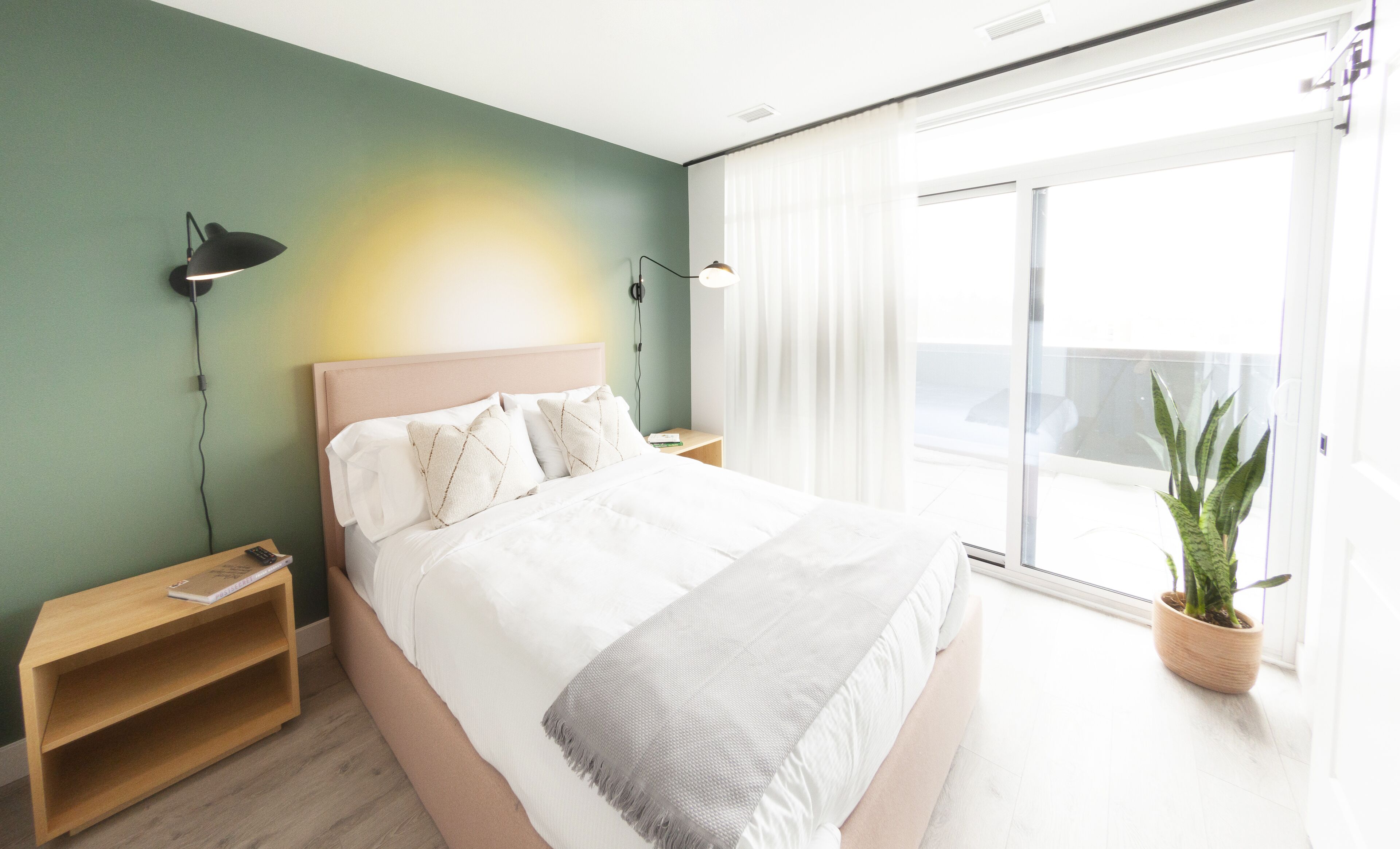 Un dormitorio moderno con una cama grande, mesitas de noche, una lámpara de noche, una planta en maceta y vistas al exterior.