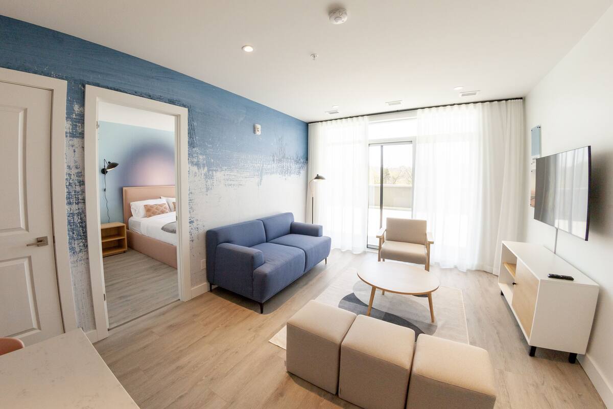 Quarto de hotel moderno com uma parede azul de destaque, um sofá, uma mesa de centro, uma TV e uma varanda com cortinas.