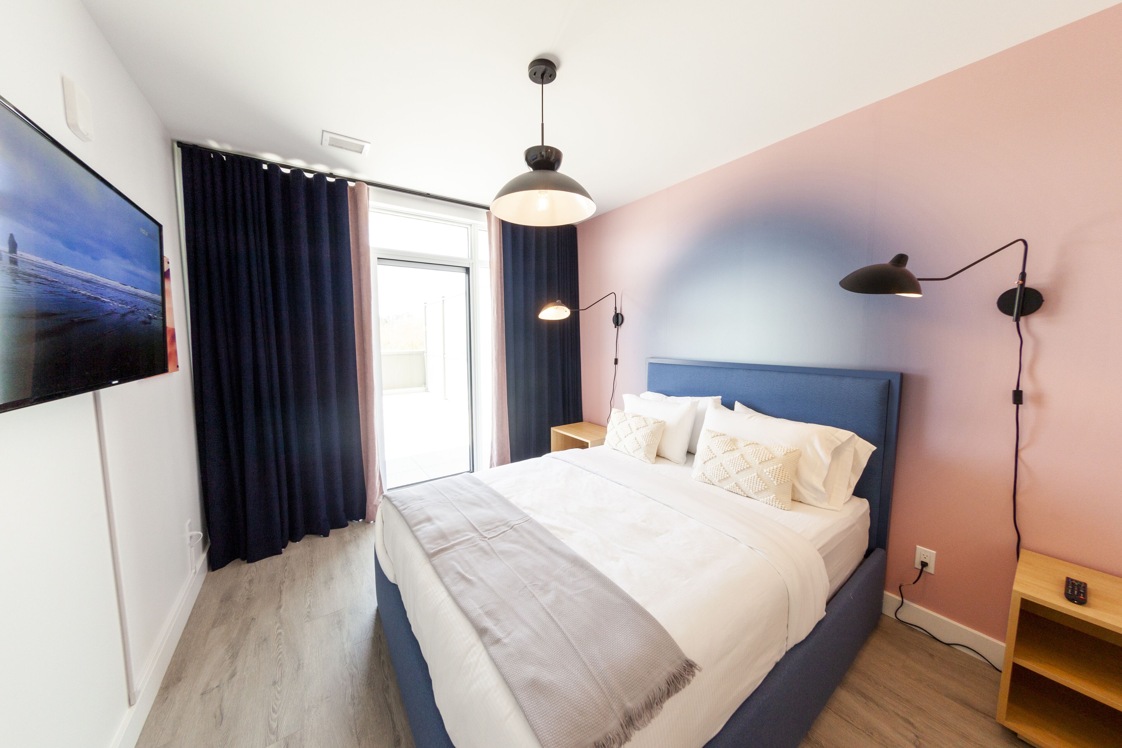 A modern bedroom with a large bed, a TV mounted on the wall, and a window with curtains.