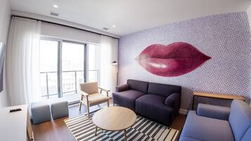 Una sala de estar moderna con un ventanal grande, un sofá, una silla, una mesita redonda pequeña y una pared decorativa con un estampado grande de labios rojos.