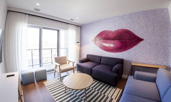 Una sala de estar moderna con un ventanal grande, un sofá, una silla, una mesita redonda pequeña y una pared decorativa con un estampado grande de labios rojos.