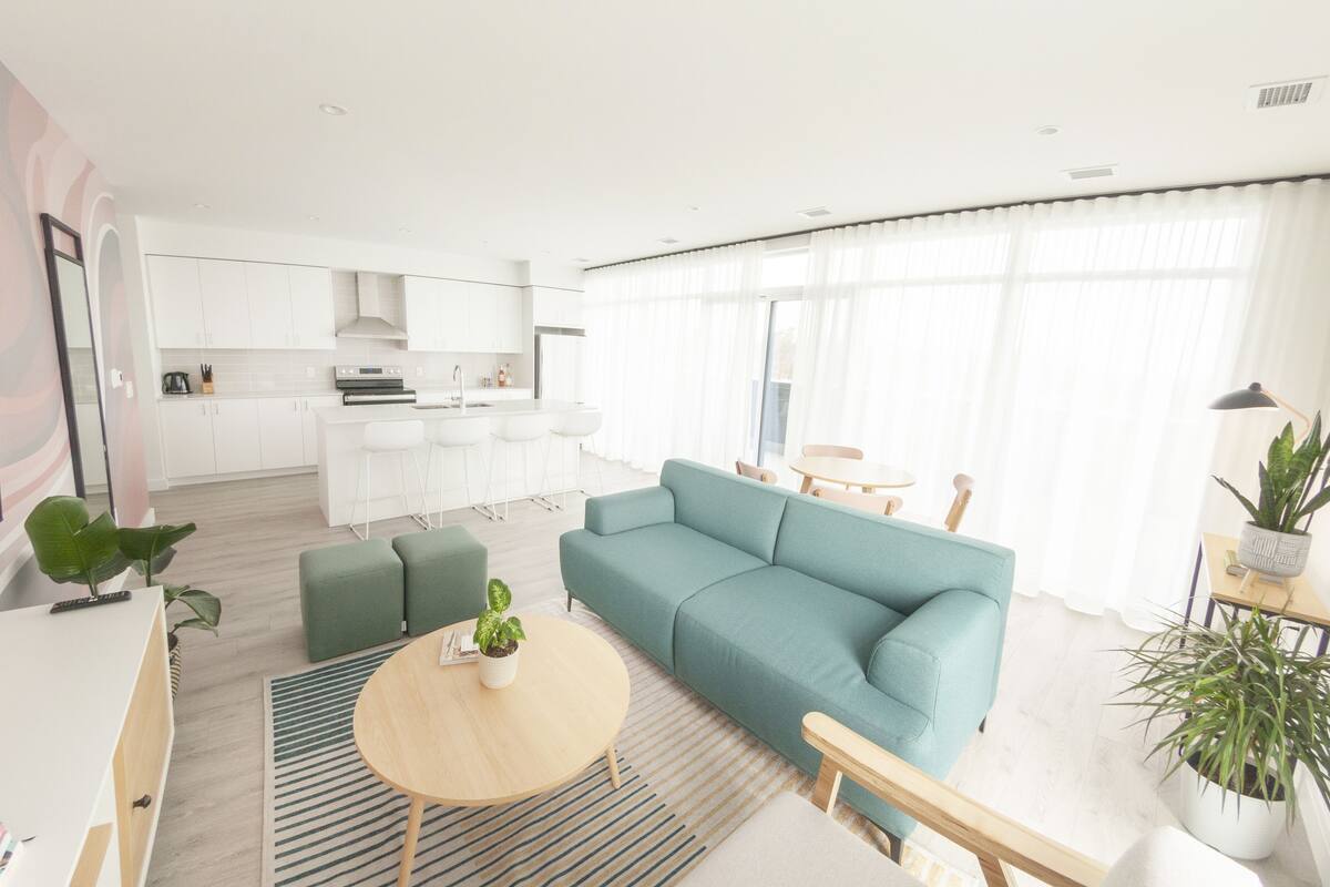 Uma sala de estar moderna com um sofá verde-água, uma mesa de centro redonda e uma cozinha ao fundo.
