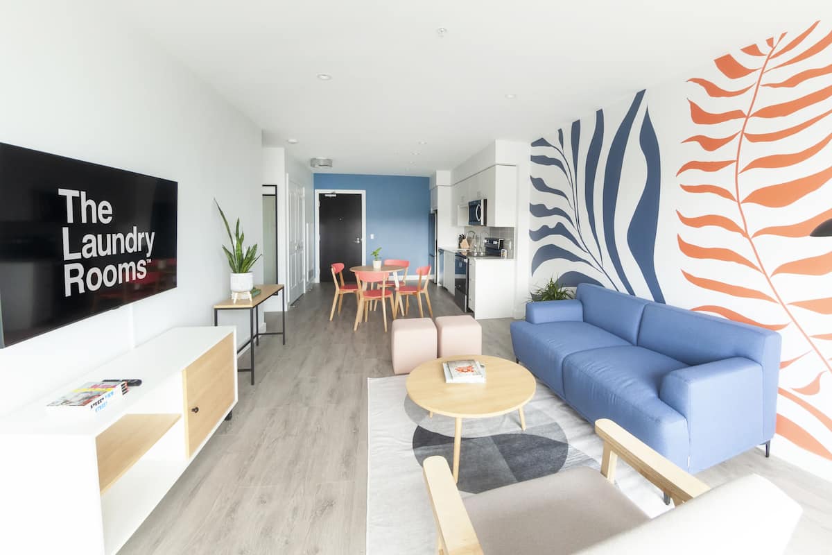 Uma sala de estar moderna, com um sofá azul, uma mesa de centro e uma área de refeições com cadeiras. Uma cozinha é visível ao fundo.
