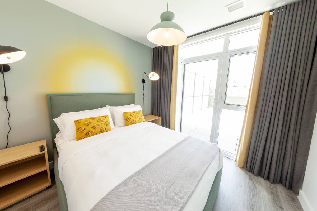 Quarto de hotel moderno com uma cama grande, mesa de cabeceira, abajur e vista para o exterior pela janela.