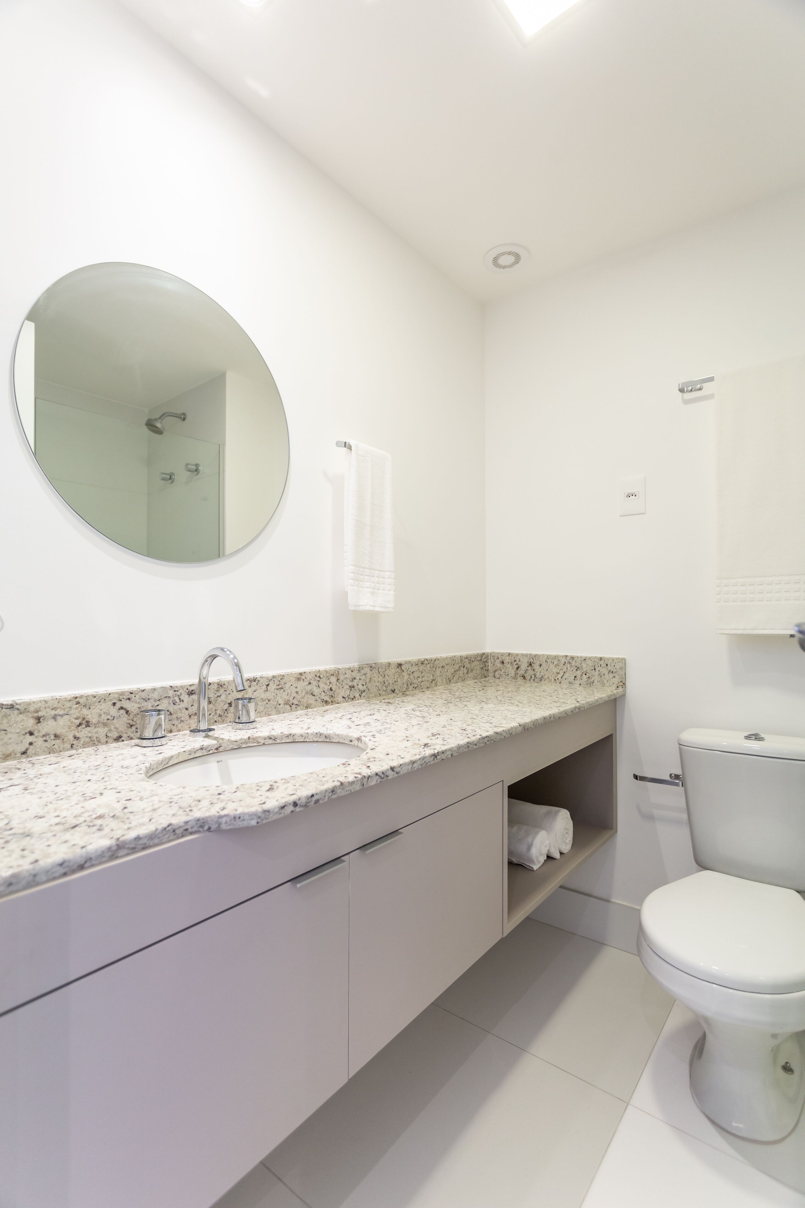 Un baño moderno con lavamanos de granito, un espejo redondo, un inodoro y un perchero para toallas.