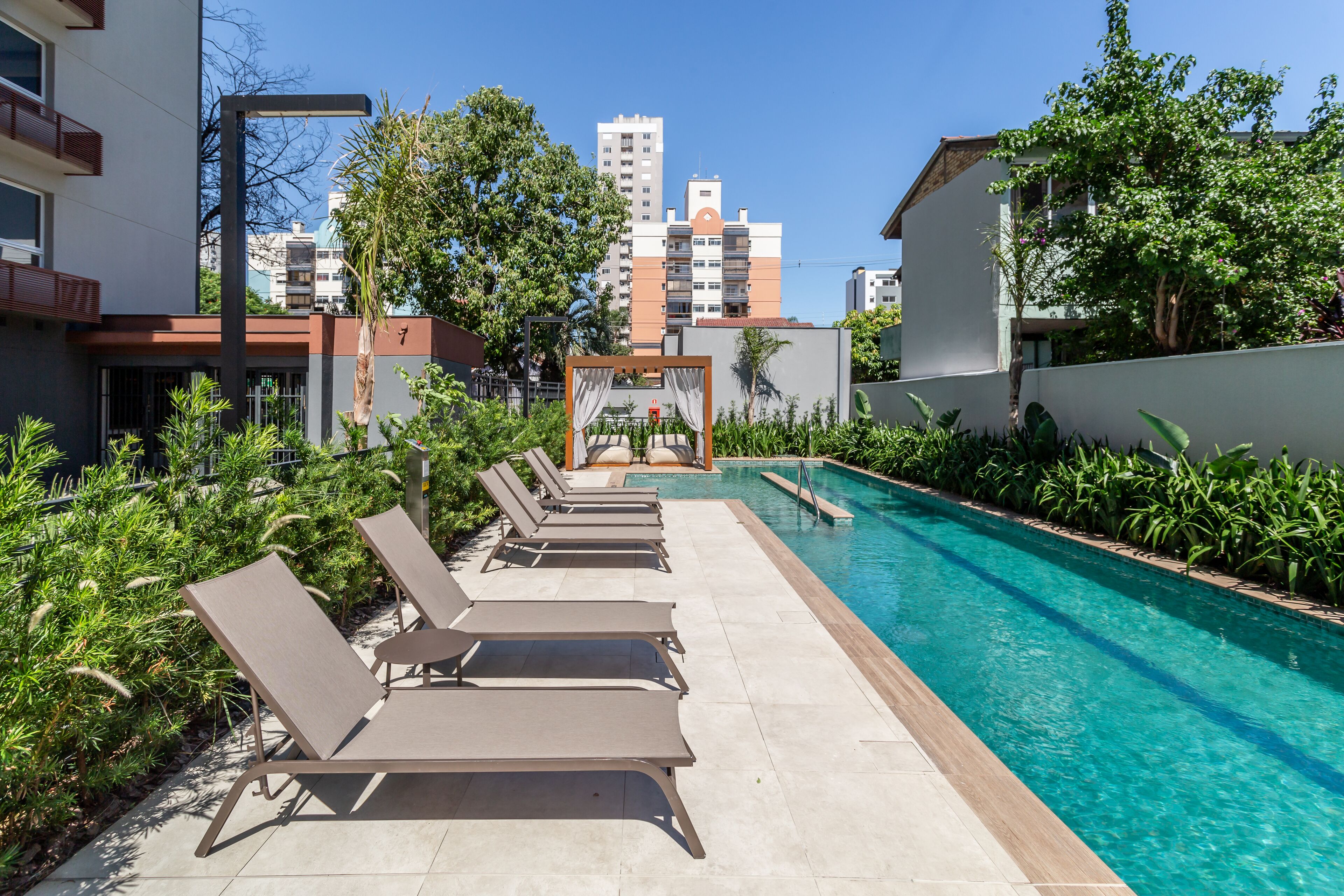 A poolside area with lounge chairs and a clear pool, surrounded by greenery and buildings in the background.