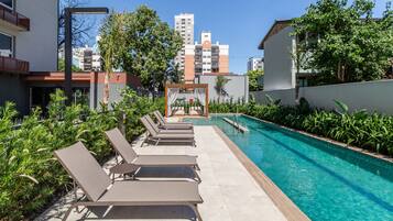 A poolside area with lounge chairs and a clear pool, surrounded by greenery and buildings in the background.