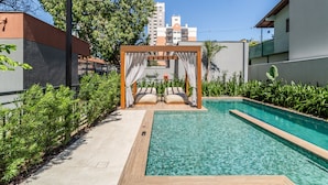 Una piscina con terraza de madera, un pabellón con cortinas y vista a un paisaje urbano al fondo.