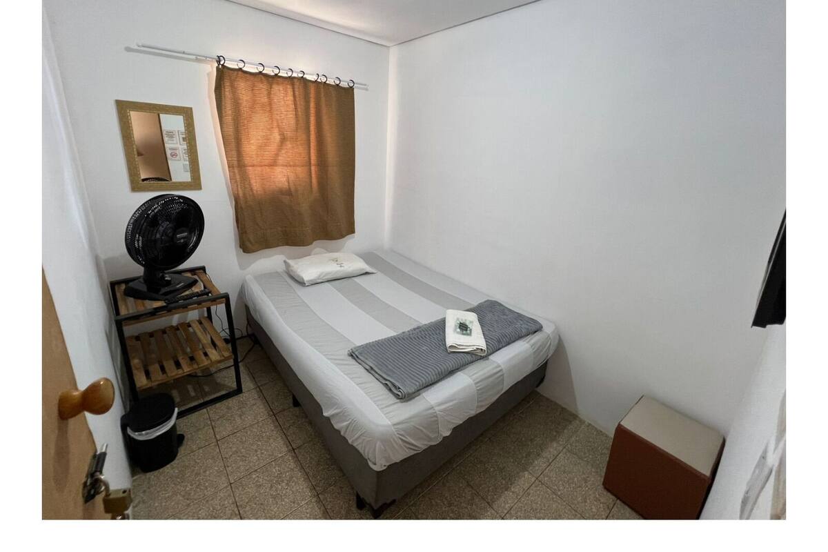 Une petite chambre propre, avec un lit simple, une table de chevet équipée d’un ventilateur, un miroir et un rideau.