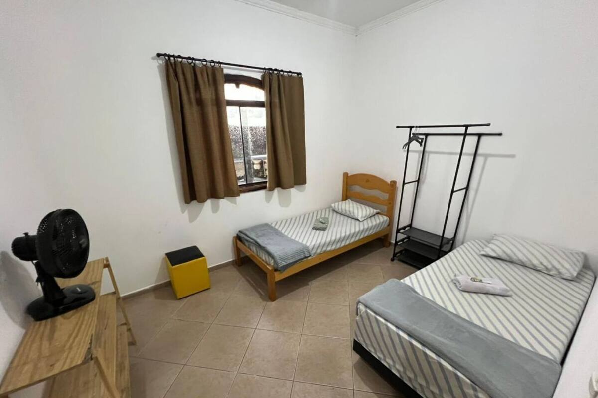 Un petit logement composé d’une seule pièce, comprenant un lit, un bureau, une chaise, un ventilateur, une fenêtre avec des rideaux et un lit superposé.