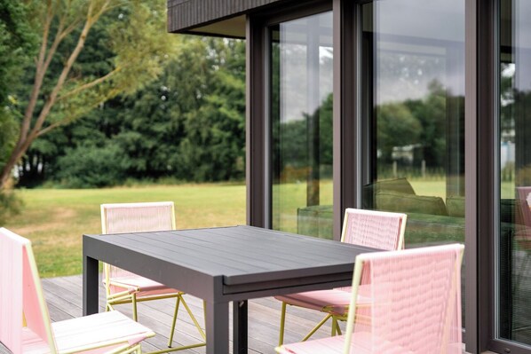 A modern outdoor patio with a dark wooden table and pink chairs, surrounded by glass walls offering a view of a green landscape. - New, top modern and stylish vacation apartment on the kl. Lake near Mirow in a dream location (Mirow)
