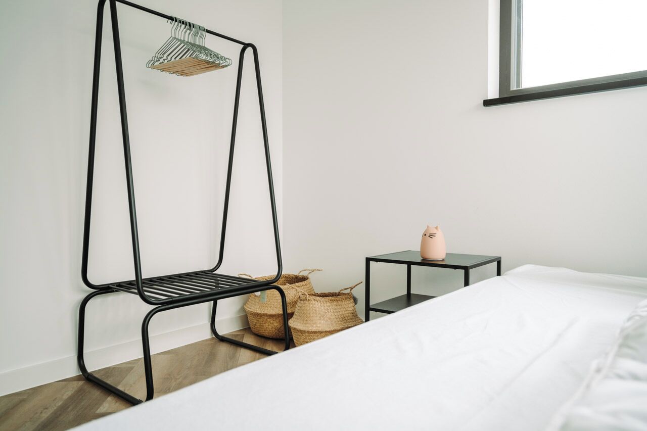 A bedroom with a bed, a black metal shelf with baskets, a small table with a pink pig figurine, and a window with blinds.
