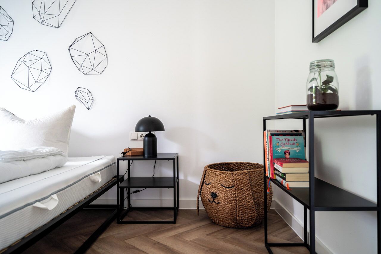 A bedroom with a bed, a bedside table with a lamp, a bookshelf, and geometric wall art.