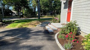 Una entrada de casa que conduce a una vivienda con puerta roja, un sendero de piedra y un jardín con flores rosas.