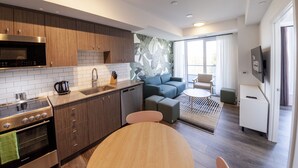 A modern kitchen with a built-in microwave, stove, and dishwasher. There is a dining area with a round table and chairs. A living area with a sofa and a TV is visible in the background. - The Laundry Rooms Station Park Kitchener (Kitchener)