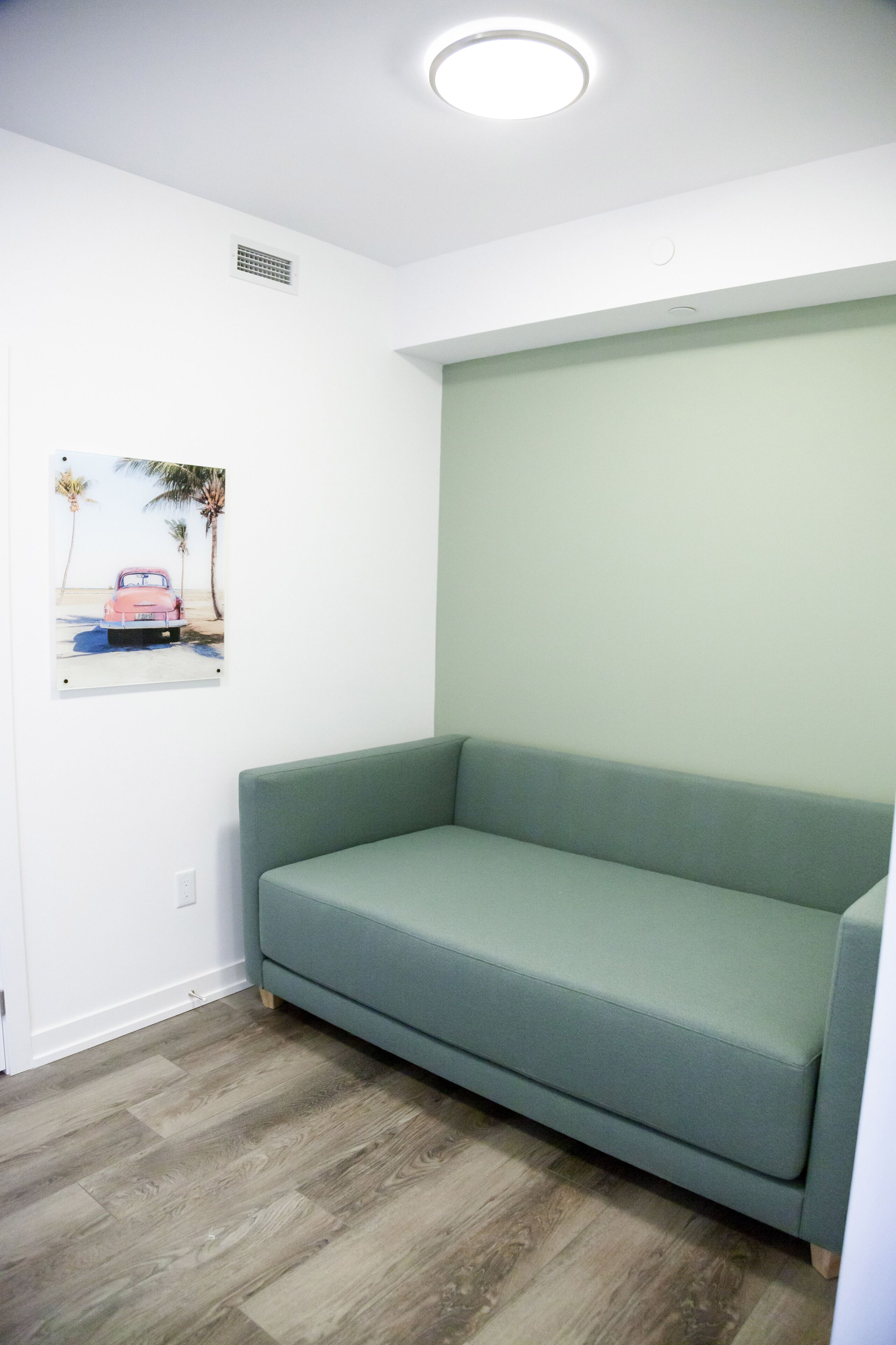 Um quarto com sofá verde, parede branca com um quadro e piso de madeira.