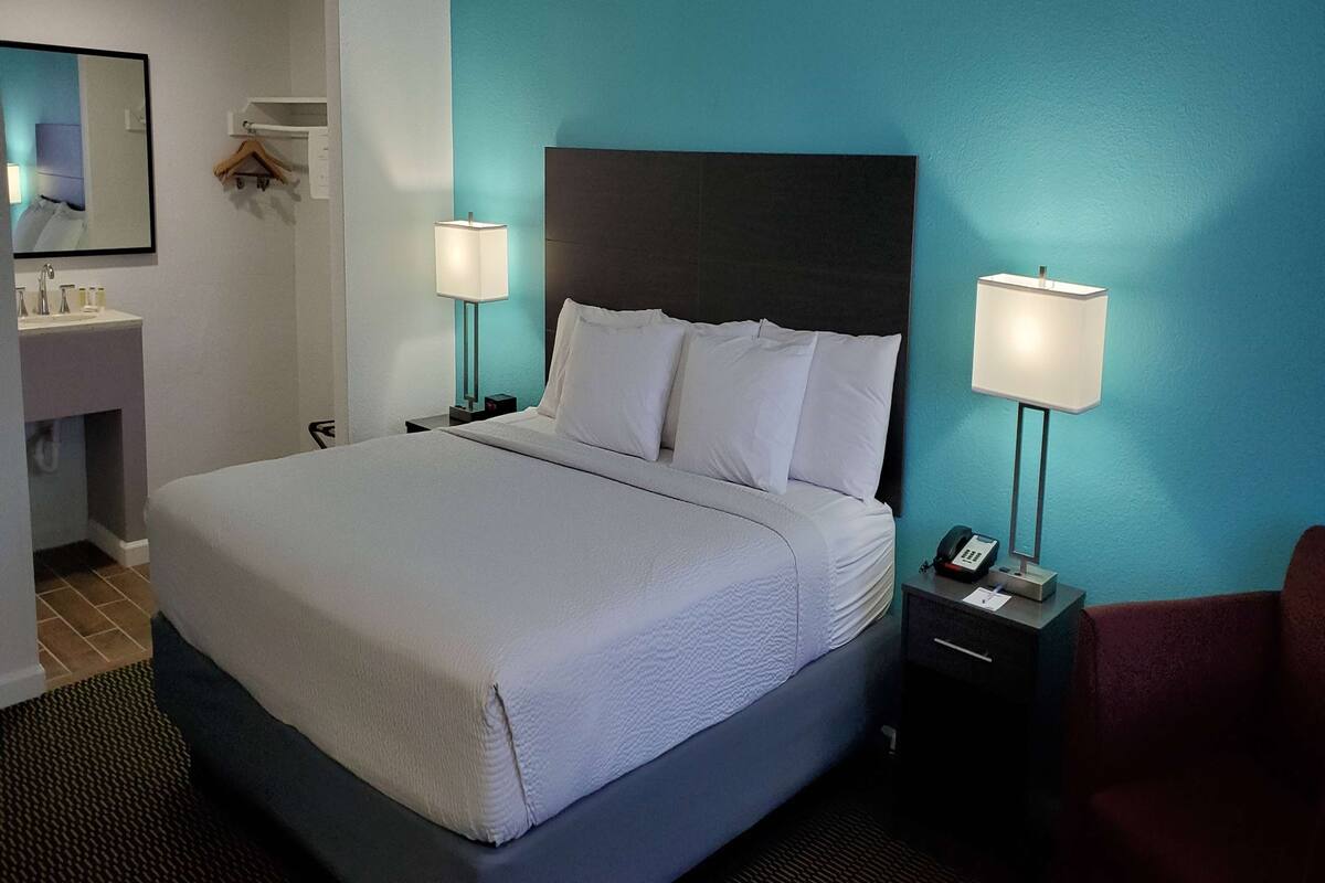 Une chambre d’hôtel avec un grand lit, deux tables de chevet avec des lampes, une petite table de chevet avec un téléphone, un miroir et une étagère murale contenant des articles de toilette.