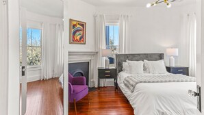 Un dormitorio con una cama grande, una chimenea, una mesita de noche, un sillón morado y una araña.