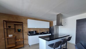Eine moderne Küche mit weißen Schränken, einer schwarzen Arbeitsplatte und einer zentralen Kochinsel mit Barhocker. Es gibt ein Holzregal mit Schüsseln und einer Kaffeemaschine.