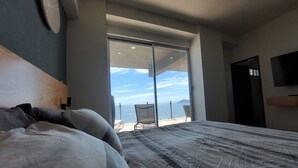 Ein Schlafzimmer mit einem großen Bett, Meerblick durch eine Schiebetür, ein Sessel und ein an der Wand befestigtes Fernsehgerät.
