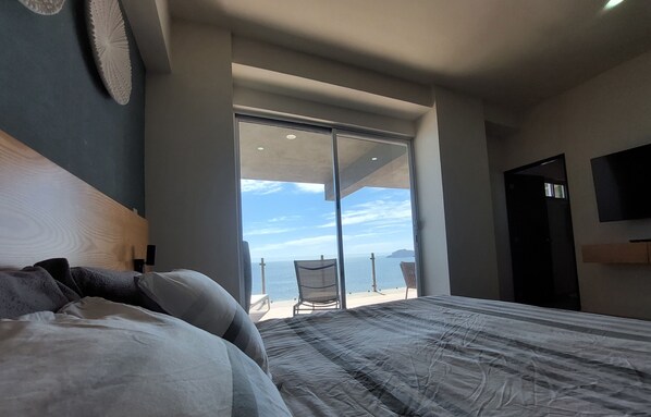 A bedroom with a large bed, a view of the sea through a sliding door, a chair, and a TV mounted on the wall.