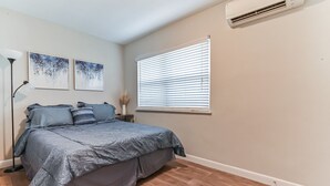 Une chambre à coucher comprenant un lit, une fenêtre avec des stores, un climatiseur et deux œuvres d’art accrochées au mur.