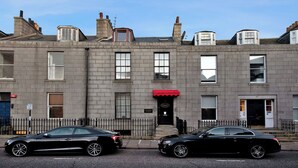 A row of grey townhouses with a red awning, black cars parked in front, and a clear blue sky.