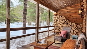 A wooden cabin with a large window, a wooden table with a tray, a chair, and a stack of firewood.