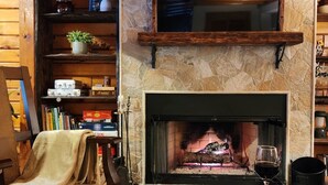 A cozy living room with a stone fireplace, wooden shelves, a comfortable chair, and a table with snacks.