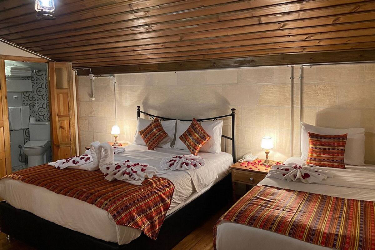 Ein Zimmer mit Holzboden, zwei Betten mit gemusterten Betttextilien, ein Nachttisch mit Lampe und ein durchgehender Tür sichtbares Badezimmer.