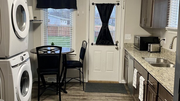 A compact kitchen with a washing machine, a dining table with chairs, a small window with curtains, and a door leading to another room.