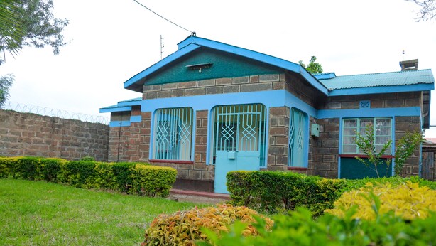 Une maison à deux étages, avec un toit bleu et des murs en briques, entourée de haies bien entretenues et d’une pelouse verdoyante.