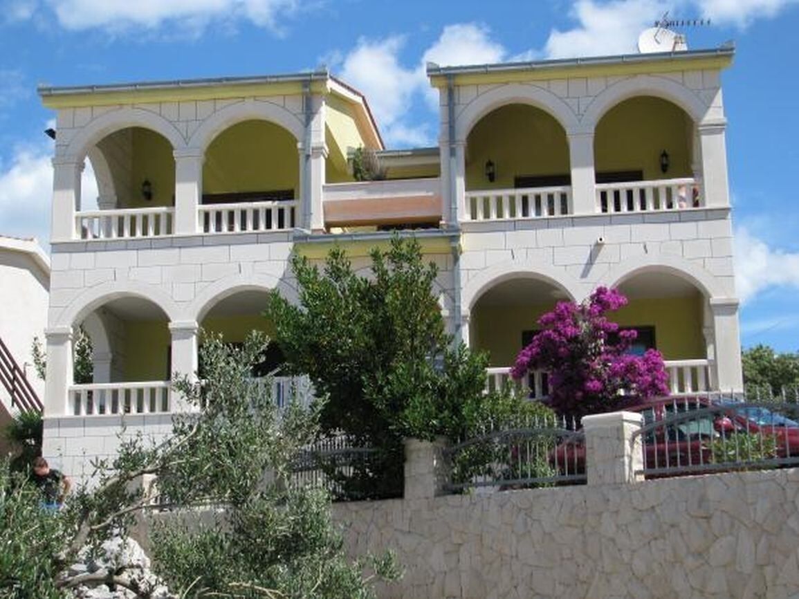 Un immeuble de deux étages avec une façade jaune, de nombreux balcons et un mur de pierre au premier plan.