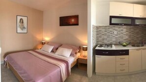 Ein Schlafzimmer mit Bett, Nachttischlampen und einem Nachttisch. Angrenzend eine Küchenzeile mit Mikrowelle, Herd und Schränken.