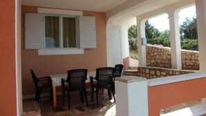 A covered outdoor seating area with a table and chairs, a window with shutters, and a stone wall in the background.